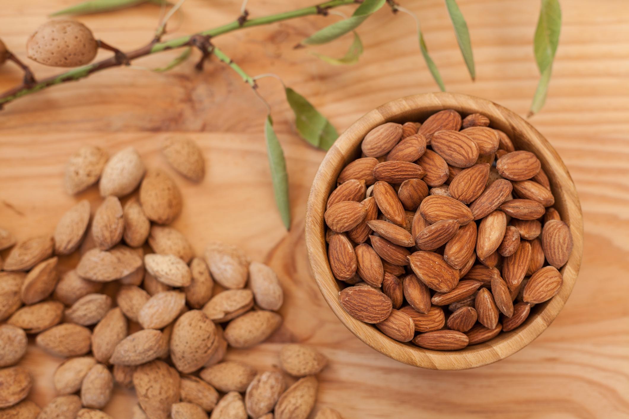Top view of shell and peeled almonds in wooden bowl and on wooden table with a unfocussed almond tree branch