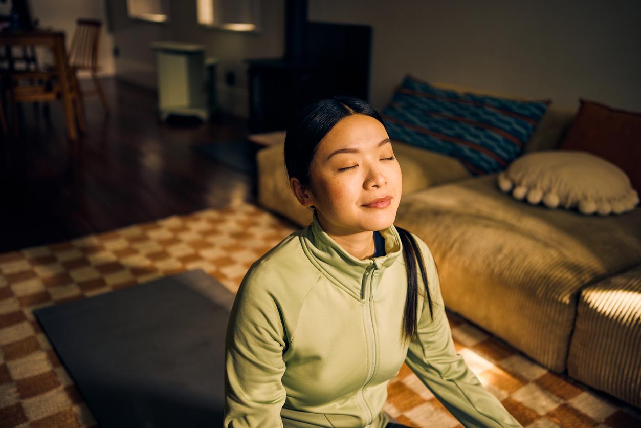 Young Asian woman meditating in living room. Indoor lifestyle portrait. Self-care and mindfulness concept.