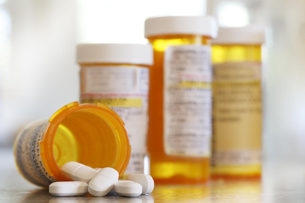 Pills pour out of a prescription medication bottle onto a kitchen counter. Several other pill bottles stand out of focus in the background.